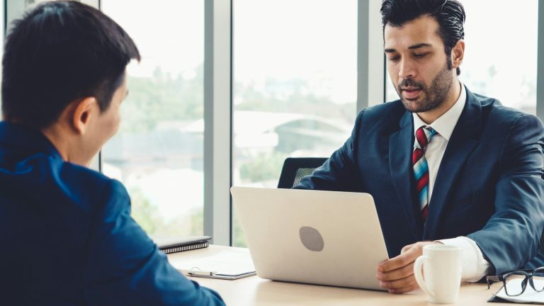 Skill Loop Collective Career Page Two men in business attire discussing over a laptop in a bright, modern office.